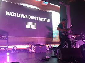 Tom Morello of Rage Against the Machine plays on a stage in front of a backdrop with the word Nazi lives don&#39;t matter