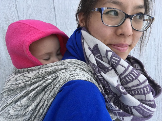 [Image of a tan skin bespectacled Asian woman looking off the frame of the image while wearing a sleeping toddler on her back who has on a pink hooded fleece jacket. Woman is wearing toddler in a gray botanical-patterned woven wrap carrier. Woman has on a dusty purple and ecru windmill-patterned infinity scarf over a blue coat. They're in front of a worn white shed.]
