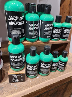 A photo of a row of cylindrical plastic clear bottles filled with bright turquoise shower gel with a black label that is rectangular that says Lord of Misrule shower gel in white font on a large light brown shelf on a bright background