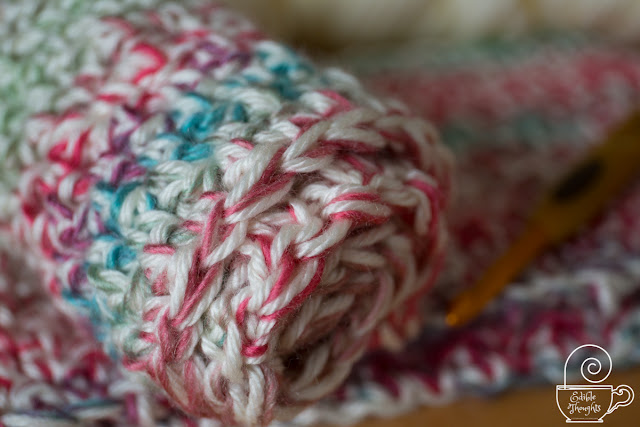 Close-up image of a multi-colored and cream yarn crocheted cowl rolled up. A golden crochet hook in out of focus on the right.