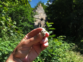 A photo of Skulferatu #43 being held up in someone's hand  and in the background are trees and the stone building Rosyth Doocot. Photo by Kevin Nosferatu for the Skulferatu Project.