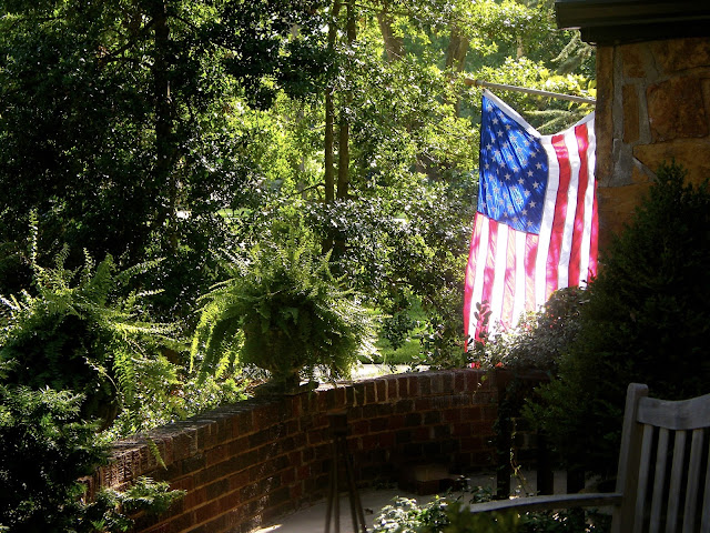 Glowing American flag is backlit by setting sun next to front door. Pots of fern grace the porch.