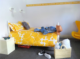 One-twelfth scale modern miniature bedroom in yellow, white and grey. On the bed is a hawaiian and baseball cap. On the floor in front is a pair of sneakers and a cane basket containing a towel, book and bottle of water.