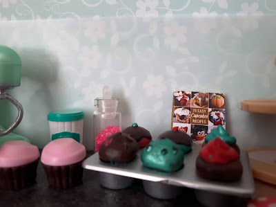 One twelfth scale modern miniature kitchen bench containing an electric mixer, a bowl with wooden spoon, several iced and decorated cupcakes, a cupcake recipe book, and a cake stand.