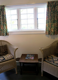 Inside view of an arts-and-crafts-style cottage, cane armchairs under a window.