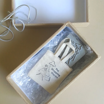 Small box on top of a padded mailing envelope, open to show a dolls house miniature tote bag and a rubber stamp.