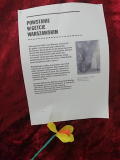 Na czerwonym suknie przypięta karta z opisem powstania w getcie warszawskim. Niżej zrobiony z papieru żonkil.