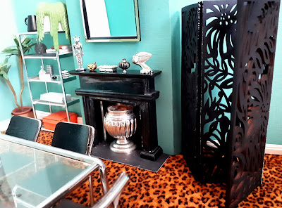 One-twelfth scale modern miniature art-deco inspired dining room with leopard-print carpet and chrome table, chairs and book shelf. To the left is a screen with a cut out flower and leaf design.