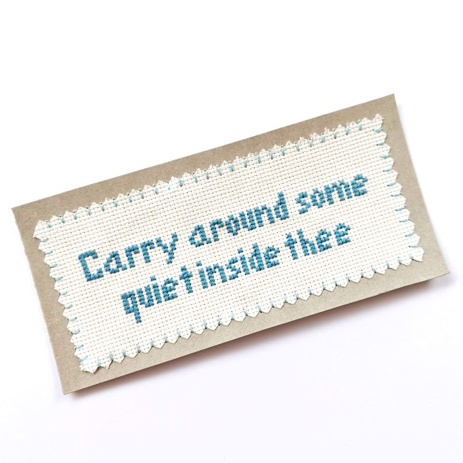 bookmark with blue cross stitched text saying carry around some quiet inside thee on aida fabric stitched around the edges on to Kraft-tex