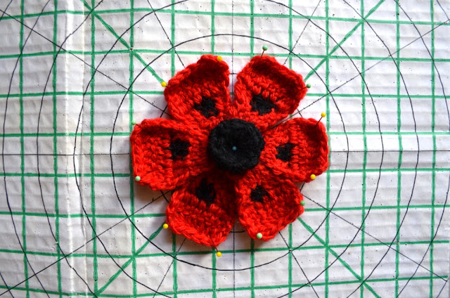 Red oriental poppy pinned to cardboard blocking board. The board is white and marked with a lined grid of squares in green ink. Two diagonal lines cross the grid at 45 degree angles. Where they intersect marks the centre of the circle. Concentric circles in dark ink radiate outwards at 1 inch spacing. These circles are broken up into cross-sections by dark lines to divide the circle into 12 sections.