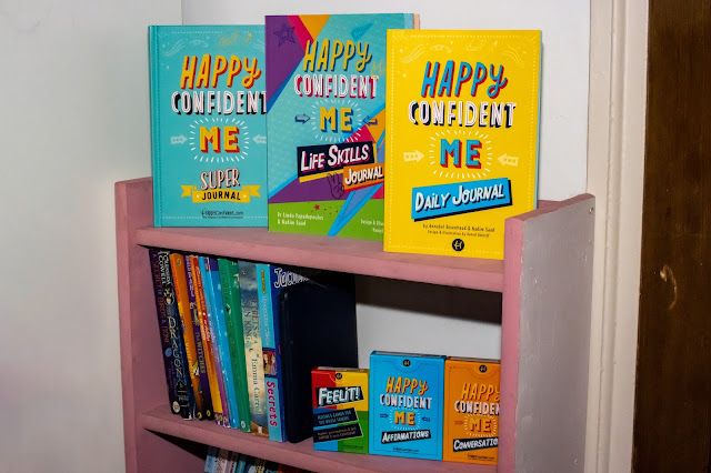 A pink book shelf in a Tween's bedroom with the Happy Confident Me collection of journals, affirmation cards, conversation cards and game cards.