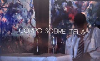 Marcos Abranches sentado a uma mesa com a cabeça baixa, garrafa e taça sobre a toalha. Na parede, dois painéis coloridos. Marcos usa camisa social branca e gravata listrada.