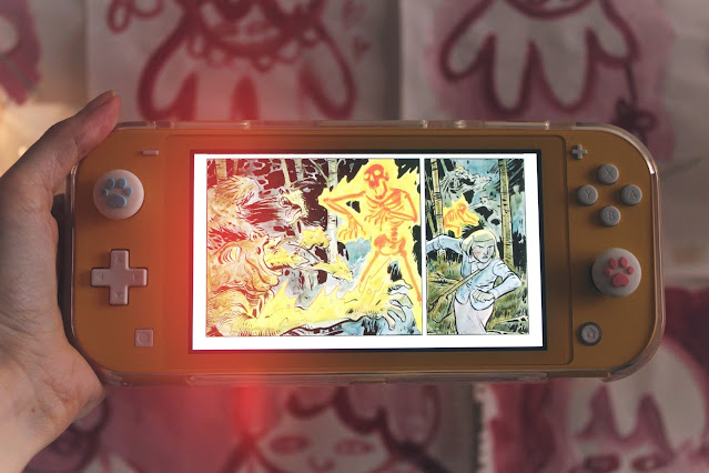 A hand holding a yellow Nintendo Switch Lite, on which a comic panel featuring a flaming skeleton can be seen.