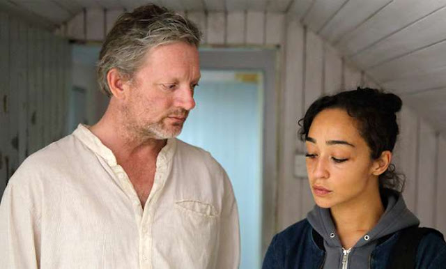 Middle-aged man looking at a younger woman in a simple, wood-planked room