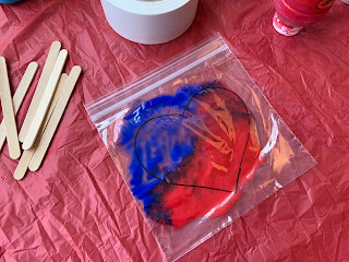 ziplock bag with heart shape filled with red and blue paint on table with popsicle sticks and duct tape