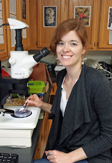 A red-haired female wearing a dark gray sweater and white blouse smiles while sitting at a microscope, holding a worm with a pair of forceps.