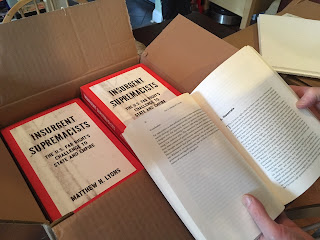 Hands holding open a copy of the book Insurgent Supremacists by a box full of copies of the book.