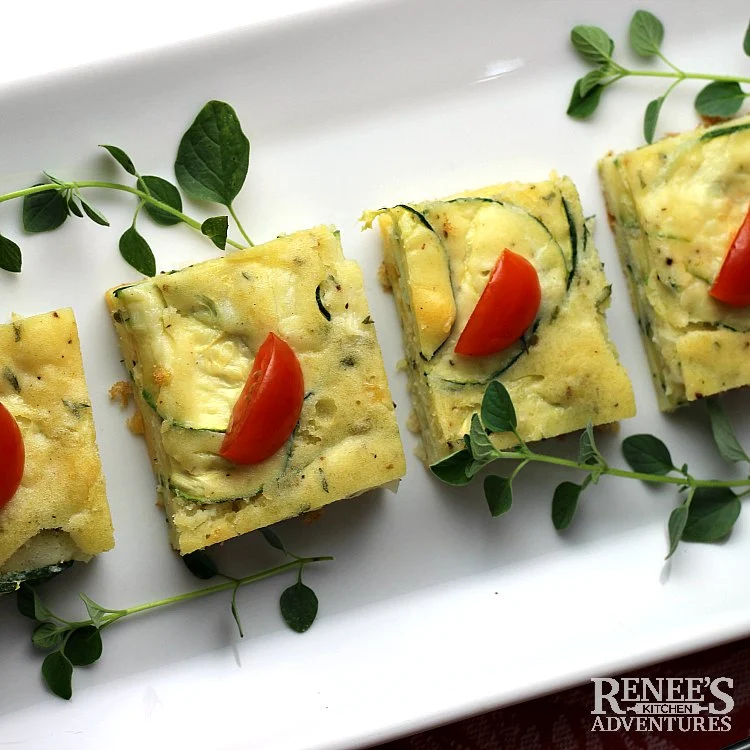 overhead shot of zucchini appetizer cut into squares on white plate with tomato and oregano garnish