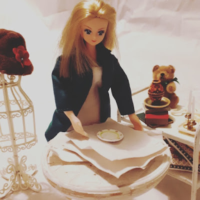 One-twelfth scale dolls' house doll behind a table with a stack of butchers paper on it and a plate on top, Behind her is a trolley containing various items including a teddy bear, a pile of books, candlesticks and cushions.
