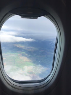 Utsikt gjennom et flyvindu over grønne jorder under noen lave skyer.