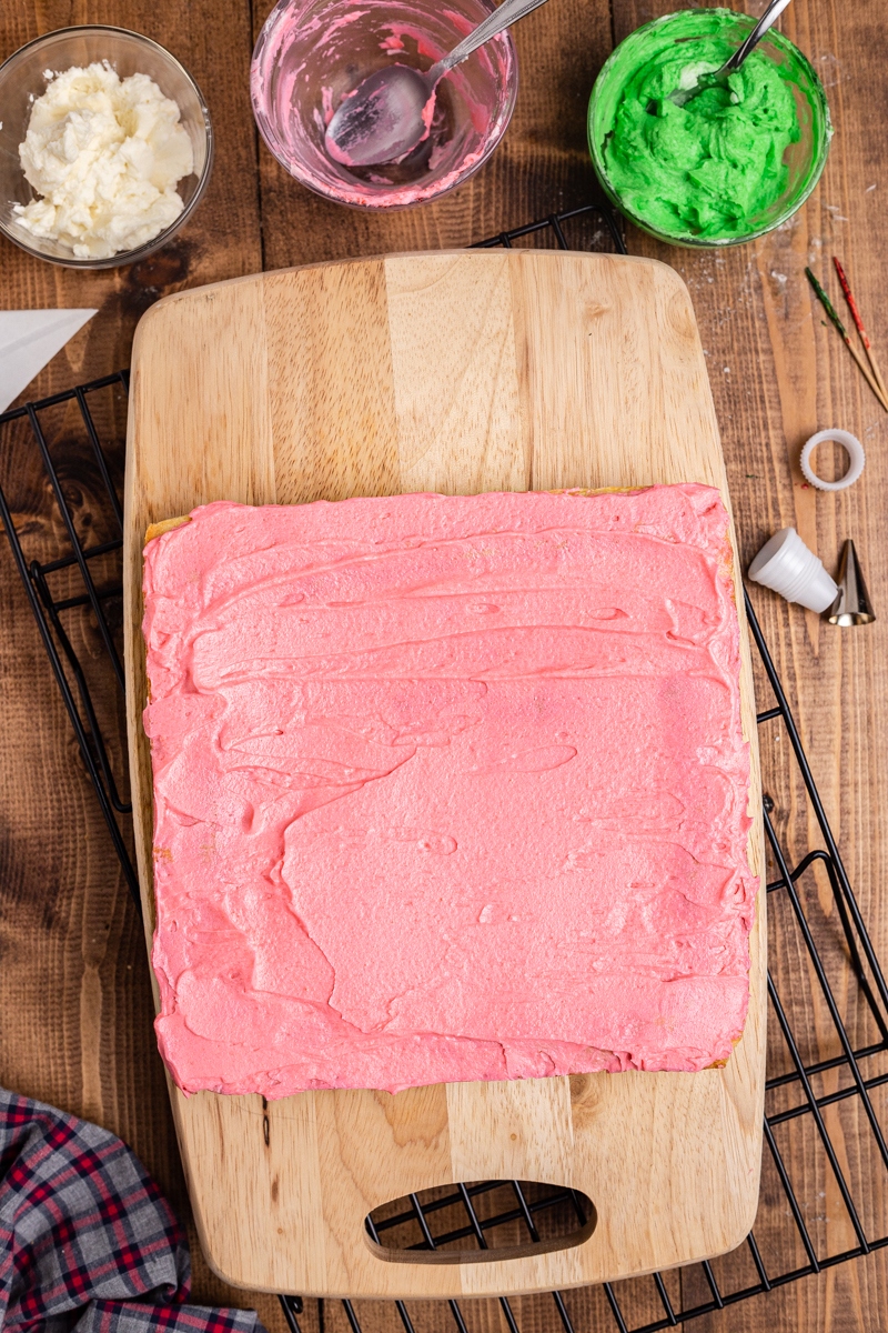 Overhead photo of Keto Sugar Cookie Bars on a cutting board covered in red frosting with bowls of green and white frosting on the side.