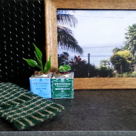One-twelfth scale modern miniature scene of a kitchen bench with two Twinings tea tins with plants growing in them, a tea towel a framed picture of a New Zealand scene displayed.