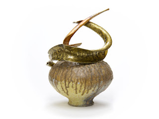 Rounded wood-fired porcelain jar with multi-coloured wood ash-glaze deposits, rests on a small foot-ring, with curved sculptural brass and bronze forms wrapped around the shoulders and top opening, viewed from the side.