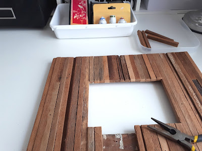 Rustic picture frame made with strips of wood. Several strips are missing and there is a set of pliers next to the missing section in the background is an Asian screen and a set of 1/12 scale ginger jars.