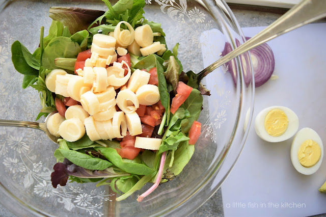 A salad is in a large, clear mixing bowl, ready to be tossed. The layers include dark leafy greens, chopped tomato, and a sliced heart palm. A sliced hard cooked egg sits beside the mixing bowl on a white cutting board. 