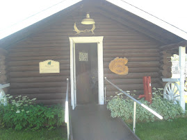 A small brown log building with a ramp leading up to a central door. Two signs are on either side of the door and a pair of antlers hang above it.