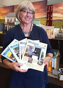 Woman standing in front of a display of one-twelfth scale miniature buildings. She is holding four copies of the Miniature Time Traveller magazine, fanned out.