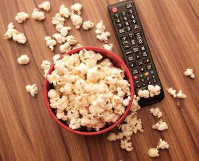 A bowl of popcorn with a lot spilled on the tabletop sits next to a tv remote.