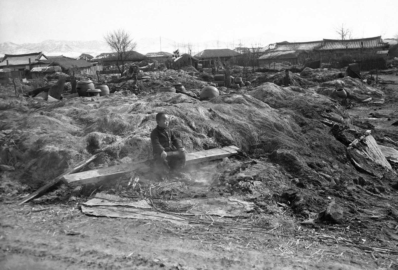 A Korean child sits in smoldering ruins of his home destroyed by fire in the Suwon area on February 3, 1951, as allied troops burned dwellings which might provide shelter for red troops. Native water jars are the only possessions recognizable in ruins of other native homes in background.