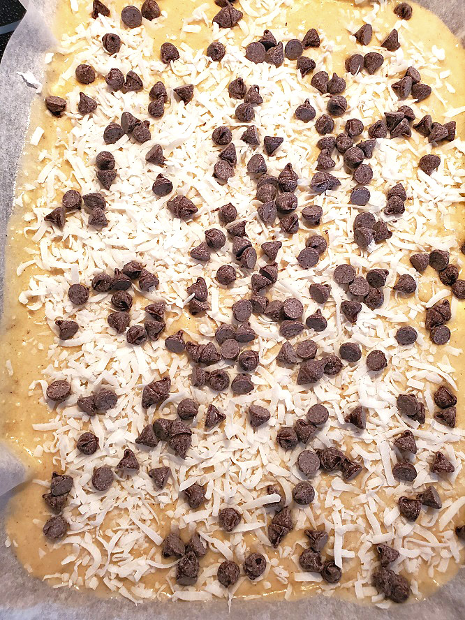 this is a pan of banana cake topped with coconut and chocolate chips in a sheet pan lined with parchment paper there are bananas in the background and pot holder on the pan