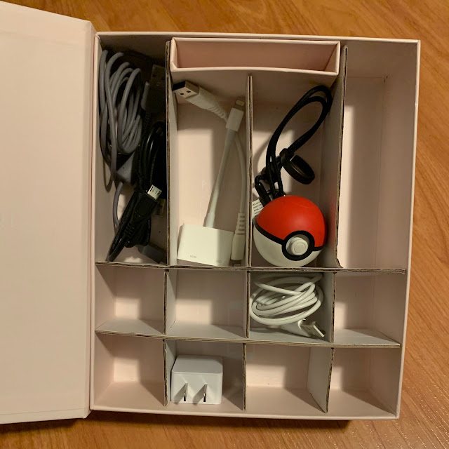 A really good box: a pink Olive and June box with a hinged lid is split into compartments by recycled cardboard strips. A Pokéball and assorted cables and chargers fill the box.