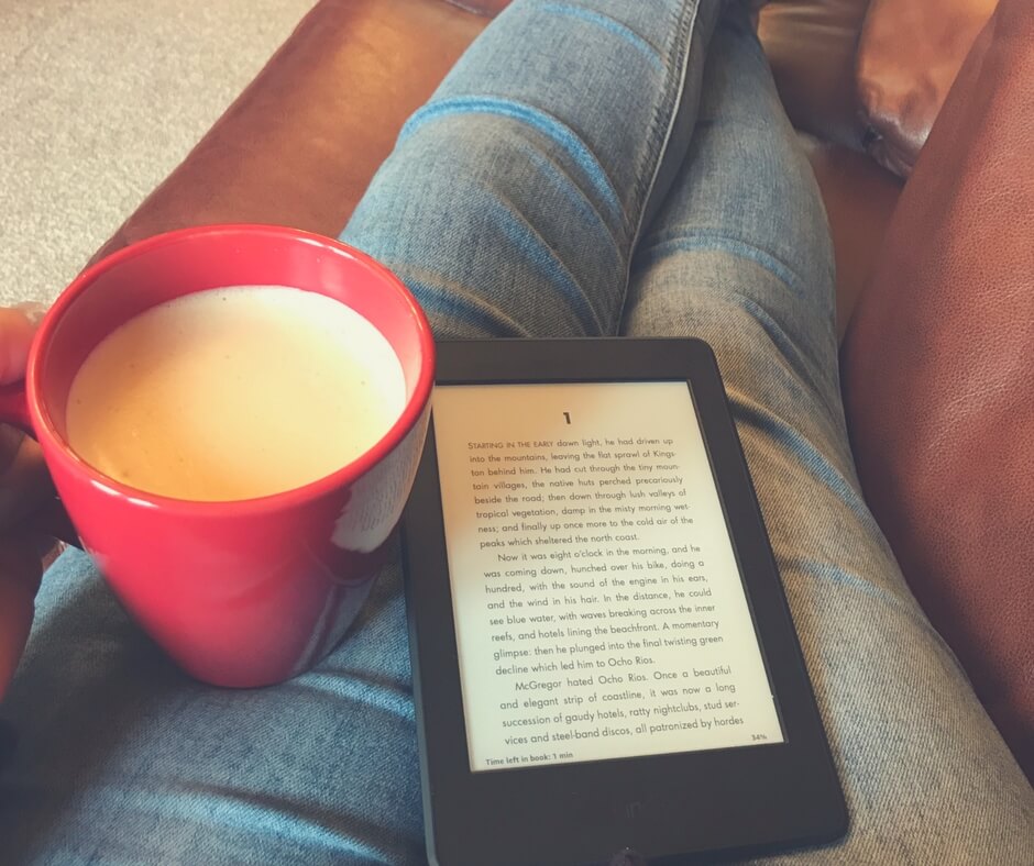 woman sits with legs up on sofa holding a red cup of coffee and reading a kindle.
