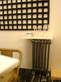 Modern dolls' house miniature half-built cafe, showing a radiator with a shelf on it, holding a water jug, glasses and a sugar bowl.