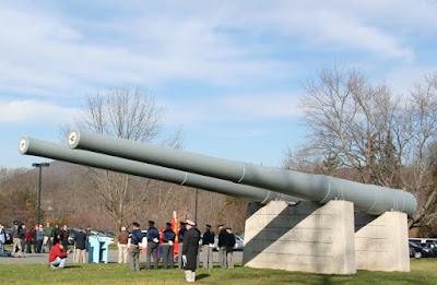 Two long gray 66-ton artillery guns from USS Pennsylvania mounted on concrete supports. Military members and civilians are gathered nearby for a public event.