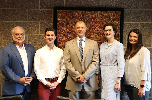 Five people, 3 men on the left and 2 women on the right, pose for the camera in front of a concrete block wall. An abstract painting hangs behind them.