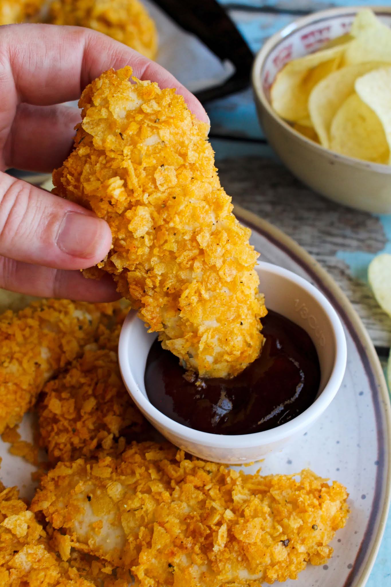 Side view of a hand dipping a Potato Chip Chicken Tender into a side of barbecue sauce.