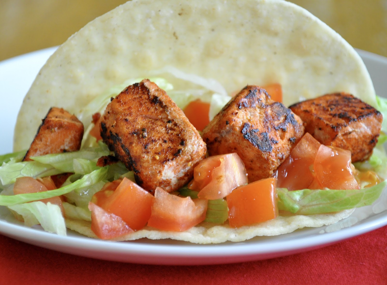 A taco featuring grilled tofu cubes, shredded lettuce, and diced tomatoes sits on a white plate, reminiscent of spicy Alaska salmon tacos in its fresh vibrancy.