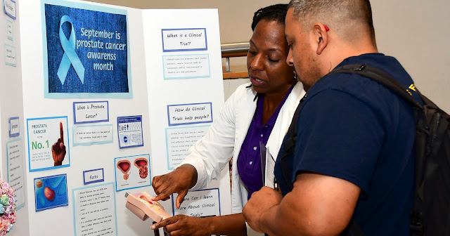A woman reads a meter to a man in front of a trifold presentation board for Prostate Cancer Awareness Month.
