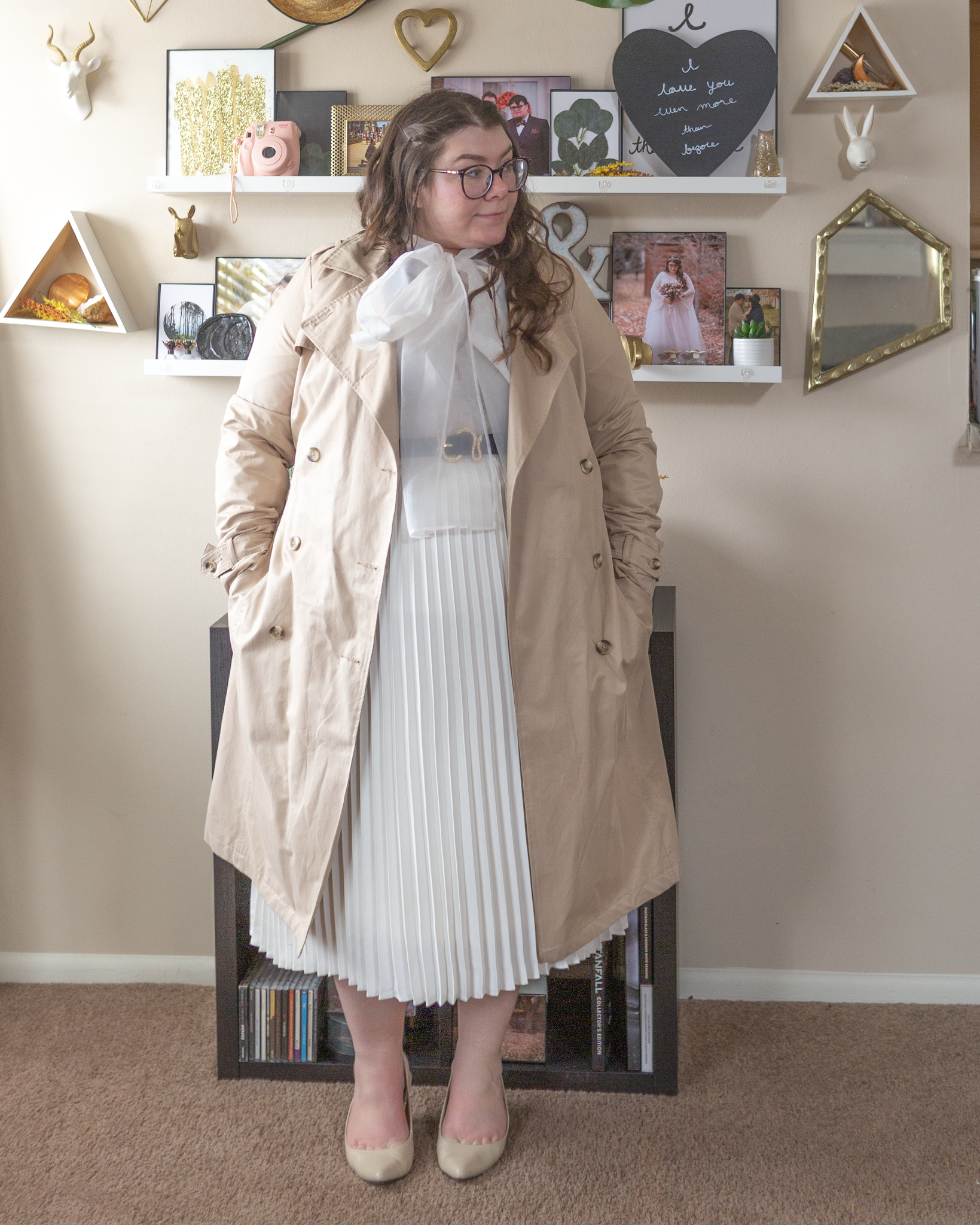 An outfit consisting of a khaki trench coat over a white sheer blouse with a large bow tied around the neck, tucked unto a white pleated midi skirt and beige slingback heels.