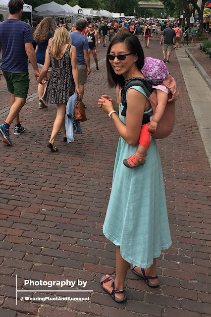 [Image of me, a light tan skin Asian woman with sunglasses, wearing a hat-donned sleeping toddler on my back with arms out in a dusty peach linen onbuhimo. I'm in a sleeveless minty colored sundress. We're standing on a brick-lined road full of people at outdoor art fair.]