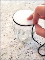 A hand holding a black piece of yarn around the circumference of a glass bottle