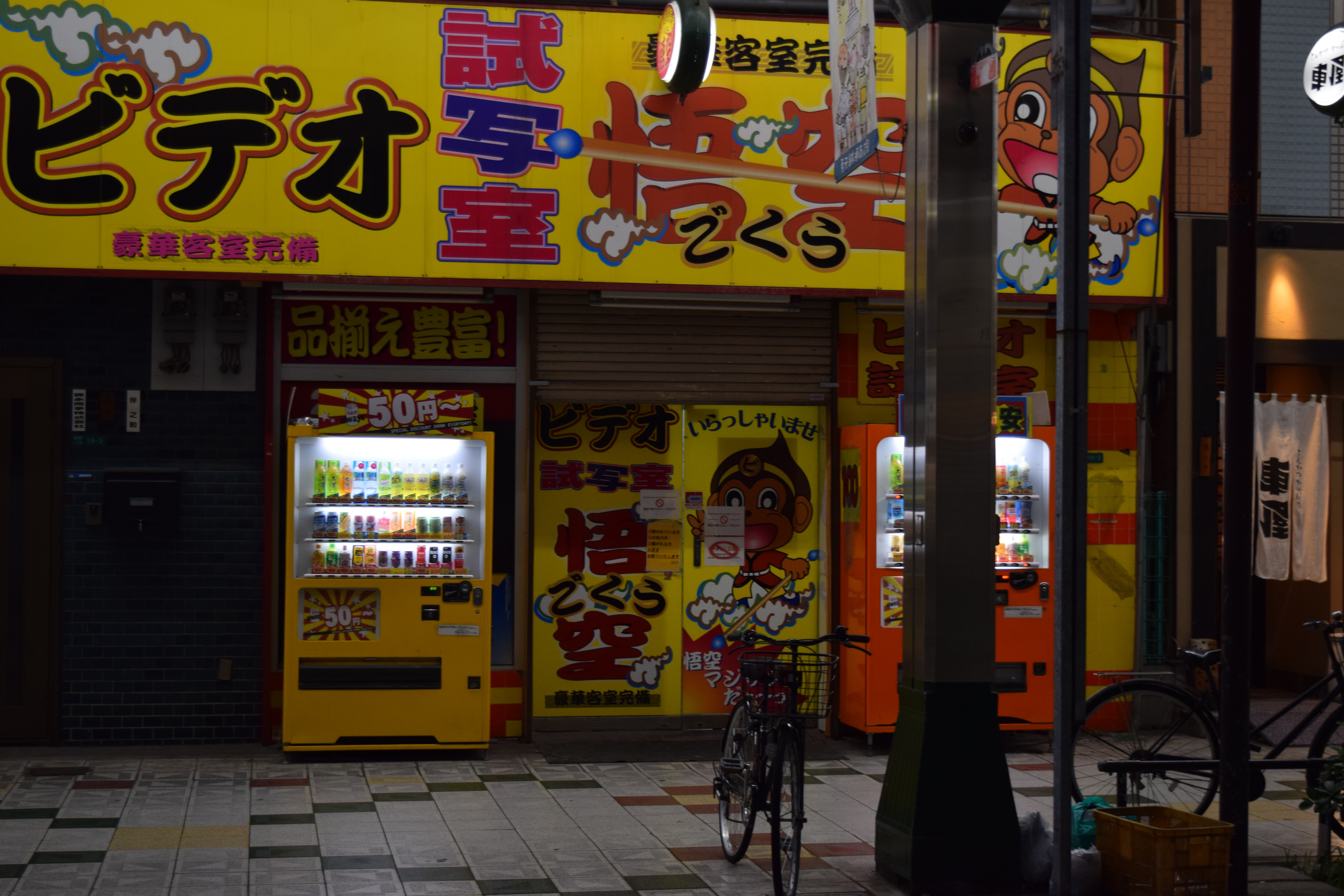 A photo of a 50 yen vending machine in Shinsekai, Osaka. It's yellow and there are lots of yellow signs in the background. There's a (mysterious) video rental shop in the background and a parked bicycle in the foreground.