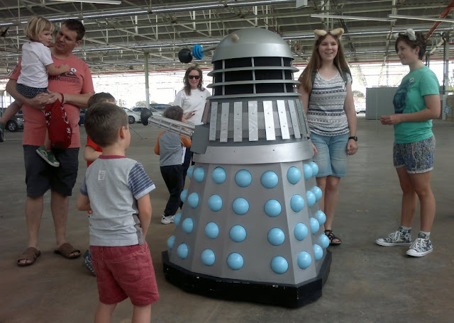 A dalek with blue details is interacting with members of the public, including children, teenagers and parents.