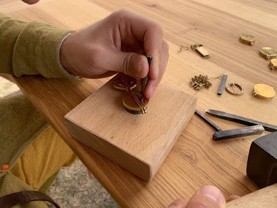 Das Bild zeigt Arme in Kleidung des 14. Jahrhunderts, die einen Metallstempel und einen Hammer halten. Unter dem Metallstempel liegt ein Spiegel auf einem kleinen runden Amboss. Der Spiegel ist zur Hälfte mit kleinen Dreiecken in geschwungenen und geraden Linien verziert.