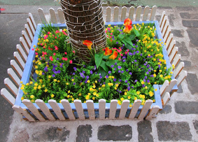 ©PatriciaYoungquist2021.This photo features a tree pit that resembles a miniature white picket fence. It is fulled with artificial flowers that are in a number of colors, including yellow, purple, blue and orange. Artificial grass is also there. It is located at Tree pit filled with artificial flowers at Lokal Mediterranean Kitchen, 473 Columbus Ave in NYC