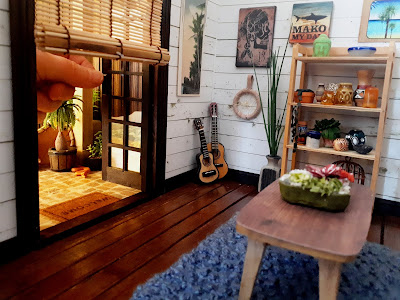 One twelfth scale modern miniature holiday house lounge with white shiplap walls and french doors opening out onto a courtyard. A full-sized hand is turning the door handle on one of the french doors.